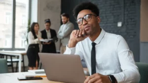 homem usando óculos sentado em frente ao laptop pensando, está em uma espécie de escritório