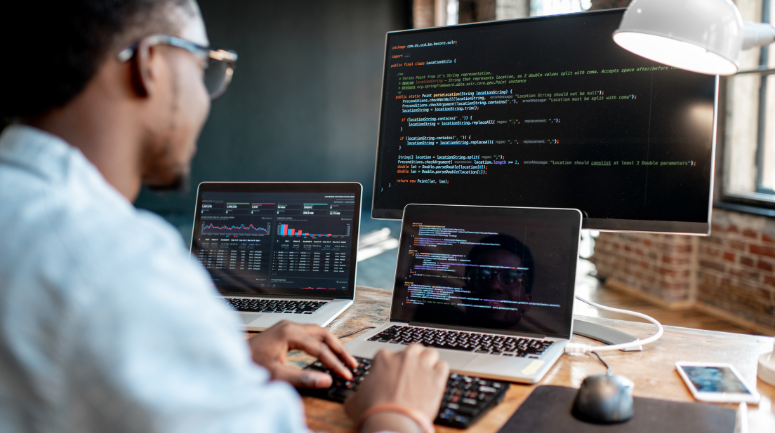 homem em frente a 3 telas de computador com códigos representando o dia do programador