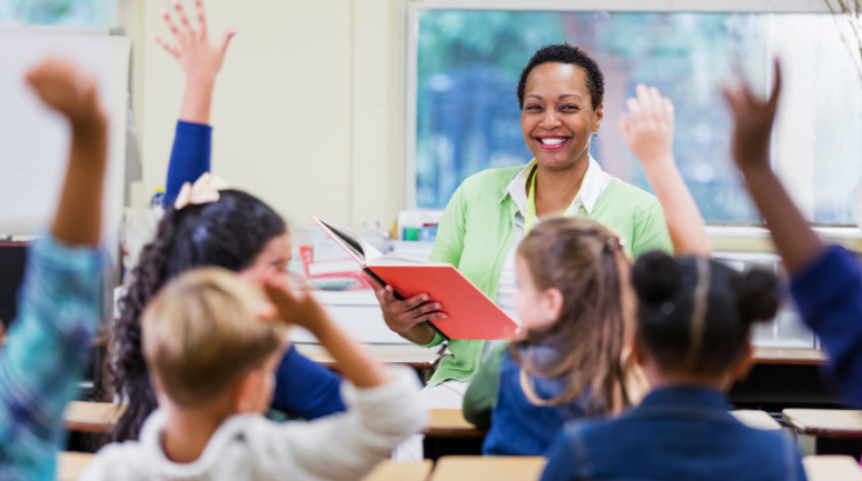 professora sorrindo com vários alunos levantando a mão na sala de aula representando dia dos professores